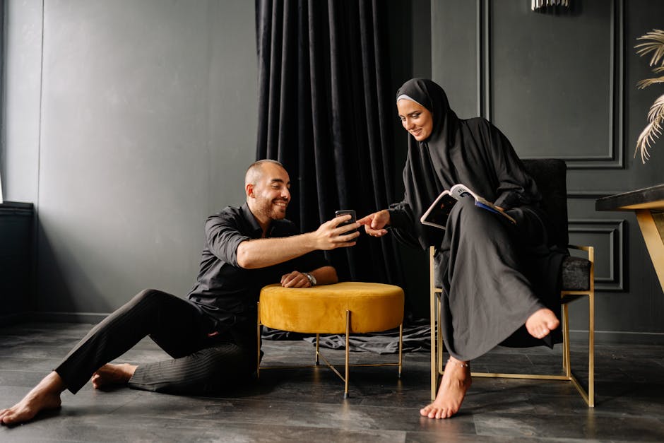 A happy couple interacting indoors while using a phone, wearing modern black outfits.